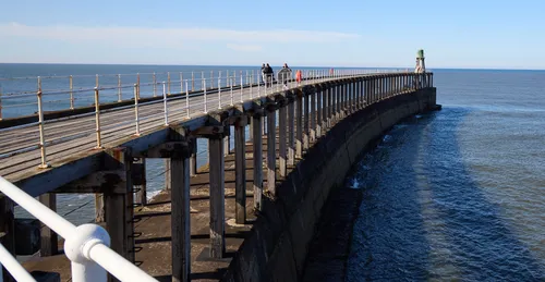 This landscape photograph captures the Whitby harbour pier extending over the water in Yorkshire. Taken during the afternoon in the spring, the image shows the pier curving gracefully out into the sea with a lighthouse visible at the end of the structure. Several people can be seen walking along the pier, enjoying the clear skies and bright sunlight characteristic of this coastal area in Whitby. The calm sea and the blue horizon emphasize the scenic beauty of the Yorkshire coastline, and the architectural details of the wooden and metal railings add a sense of history to the scene.